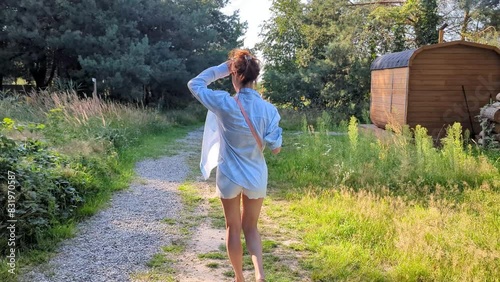 A personal small hotel sauna. Mobile compact bath in the form of a wooden barrel. SPA, relax. Back view of the caucasian woman walking in forest