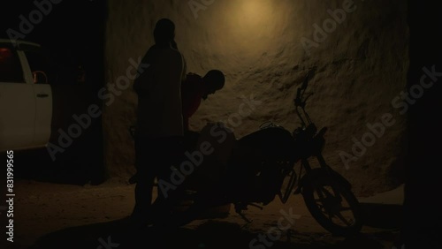 Two african men remove large tank from motorcycle near wall of house late in evening. Hard daily life in slums