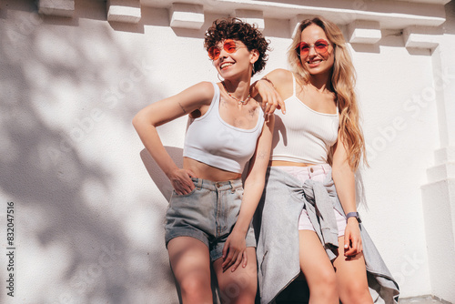 Two young beautiful smiling hipster female in trendy summer white t-shirt and shorts clothes. Sexy carefree women posing in the street. Positive models having fun, hugging and going crazy, near wall