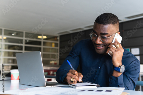 Handsome african manager sitting at office desk in front of laptop hold mobile phone make pleasant business or informal call. Successful businessman looking at financial statistic shown on documents