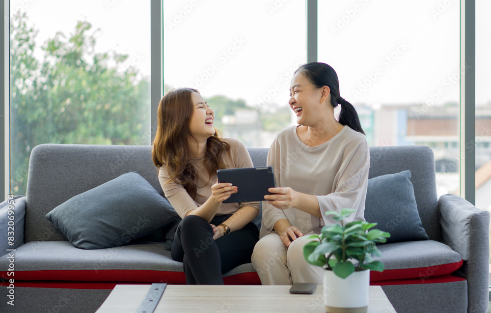 Two women are sitting on a couch, laughing together and sharing a tablet computer, in a bright living room. Two women are sitting on a couch, laughing together and sharing a tablet computer, in a bright living room.