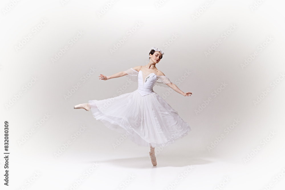 Portrait of graceful ballerina in flowing white dress, looks as fantastic character performing ballet move on pointe against white studio background. Concept of art, fusion of classic and modernity.