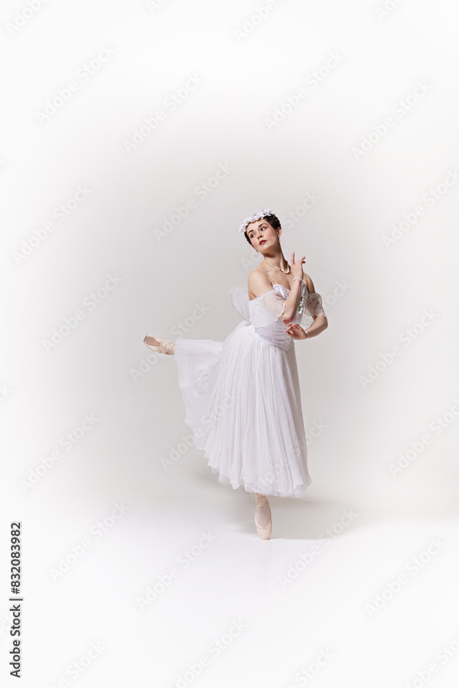 Young woman, ballerina in flowing white dress performs ballet move, conveying lightness and grace against white studio background. Concept of art, fusion of classic and modernity, grace and elegance. Young woman, ballerina in flowing white dress performs ballet move, conveying lightness and grace against white studio background. Concept of art, fusion of classic and modernity, grace and elegance.