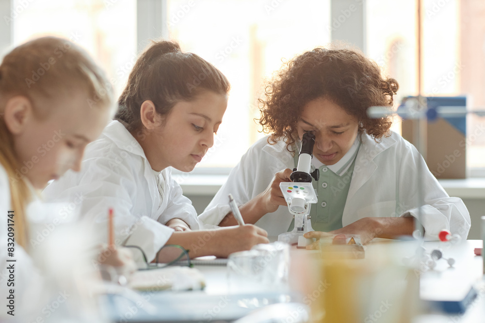 Selective focus shot of ethnically diverse middle school students wearing lab coats using microscope and making note during Chemistry lesson Selective focus shot of ethnically diverse middle school students wearing lab coats using microscope and making note during Chemistry lesson