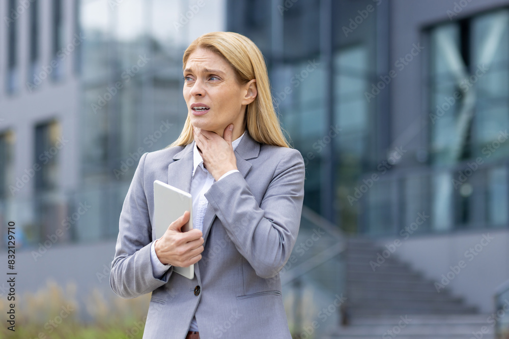 Fototapeta premium Businesswoman in a gray suit clutching her throat in discomfort while holding a tablet outside an office building.