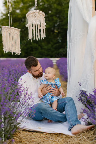 Bearded man in white shirt is sitting on a white blanket and playing with his little daughter in a lavender field. Dad with a baby. Lifestyle. 