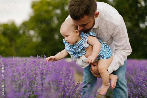A father is holding and showing a baby lavender flowers in a lavender field. A little baby in the father's arms is touching a lavender flower in the field. Baby is exploring the world. 