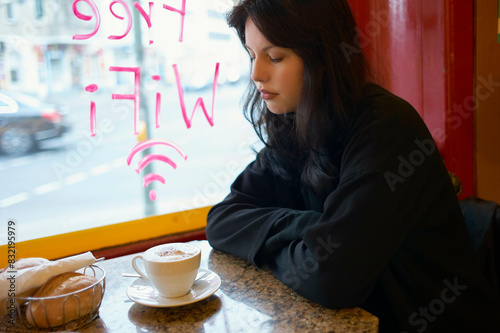 A woman sits contemplatively by a window in a cafe, with a cup of cappuccino and a sandwich on the table. photo