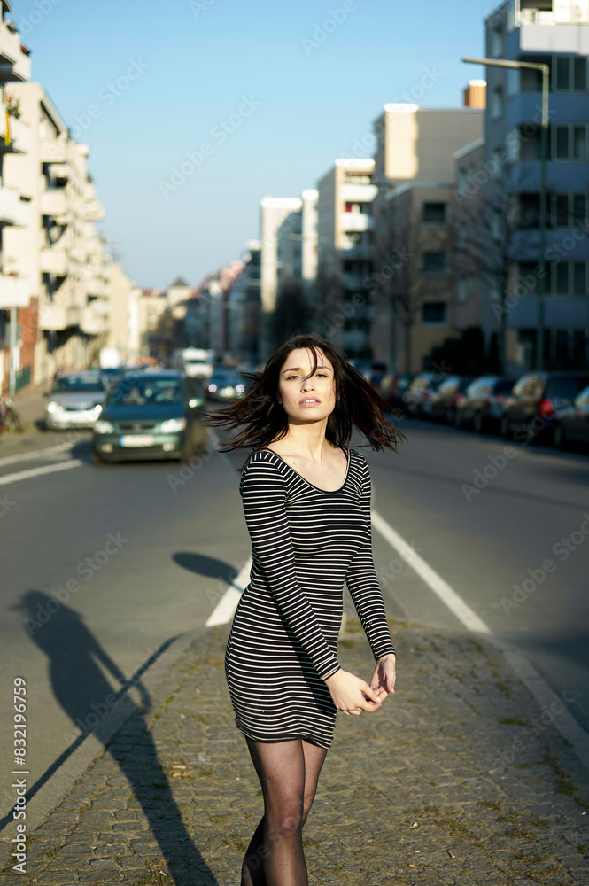 A woman walks on a sunny street with buildings and parked cars in the background. She wears a striped dress and the sun creates a long shadow on the pavement. A woman walks on a sunny street with buildings and parked cars in the background. She wears a striped dress and the sun creates a long shadow on the pavement.