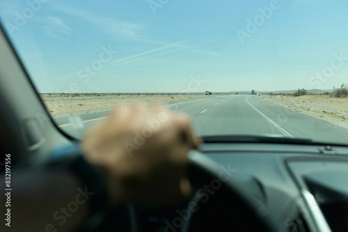 A driver's hand on the steering wheel seen from the interior of a car, with a clear view of a straight road ahead and a blue sky. photo