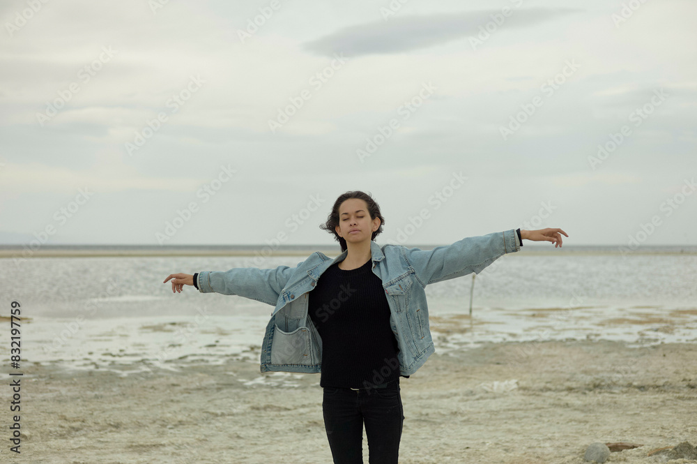 A person stands on a sandy shore, arms extended to the sides, with a cloudy sky overhead. A person stands on a sandy shore, arms extended to the sides, with a cloudy sky overhead.