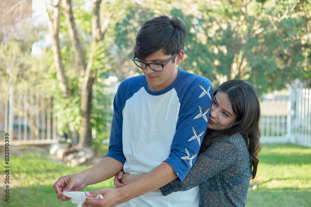 A young adult male and female are closely engaging with each other outdoors, with the female embracing the male from behind while they both look down at an object in the male's hands. A young adult male and female are closely engaging with each other outdoors, with the female embracing the male from behind while they both look down at an object in the male's hands.