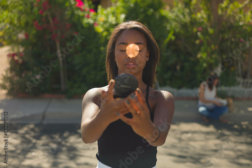 A focused young woman holds a camera lens up to her eye, taking a photo, with another person blurred in the background sitting on a bench surrounded by green foliage. photo