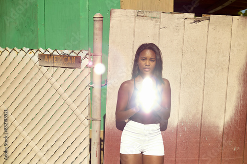 A woman stands in front of a rustic backdrop holding a bright light source centered in front of her, which illuminates the scene.