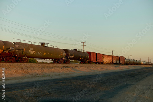 A freight train composed of various tank and boxcars is stationary on the tracks during a calm twilight, with a clear sky above.