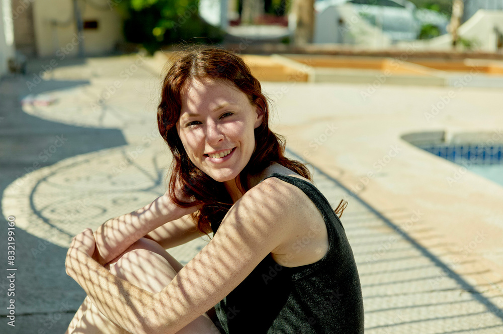 A smiling woman with red hair sitting outdoors with a swimming pool and palm trees in the background. A smiling woman with red hair sitting outdoors with a swimming pool and palm trees in the background.