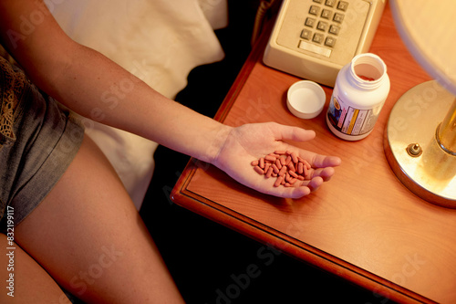 A person is sitting beside a bed with an open prescription bottle and a hand full of pills, indicating they are about to take medication.