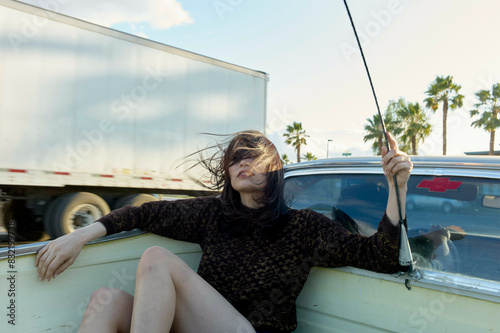 A woman is seated in a classic white convertible car holding a radio antenna with her hair blowing in the wind, with palm trees and a passing truck in the background. photo