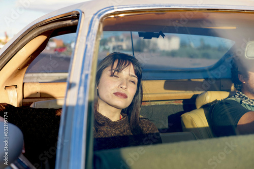 A woman with dark hair is seated in the front passenger seat of a vintage car, gazing out of the window with a thoughtful expression as the driver is partially visible with focus on the woman. photo