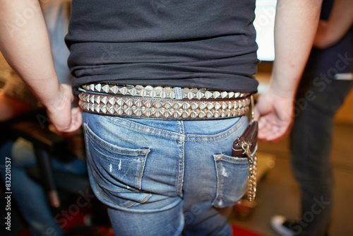 A close-up view of a person wearing blue denim jeans and a black shirt tucked in, with a studded belt and keychain accessory attached to a belt loop.