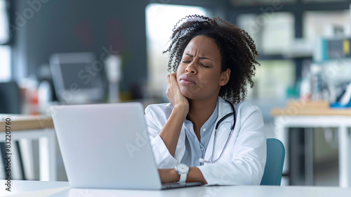 An African American woman doctor, experiencing neck discomfort and looking frustrated, reflecting the effects of hospital burnout and overwork, with a laptop on her desk