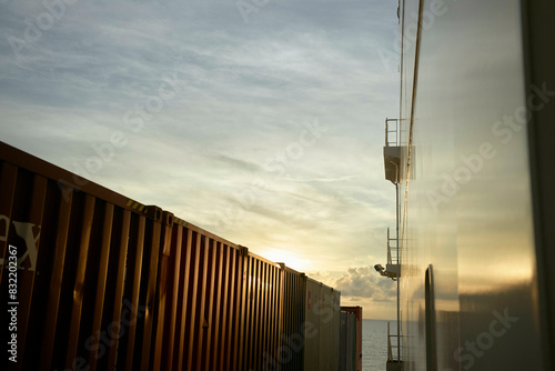 The image captures the side of a cargo ship with stacked shipping containers at sunset, with the sun dipping near the horizon, casting warm light and soft shadows. photo