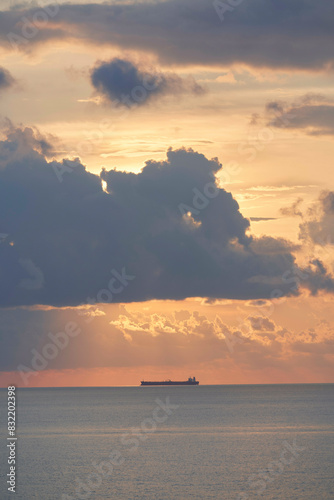 A cargo ship is silhouetted against the vibrant orange hues of a sunset sky, with large clouds partially obscuring the sun. photo