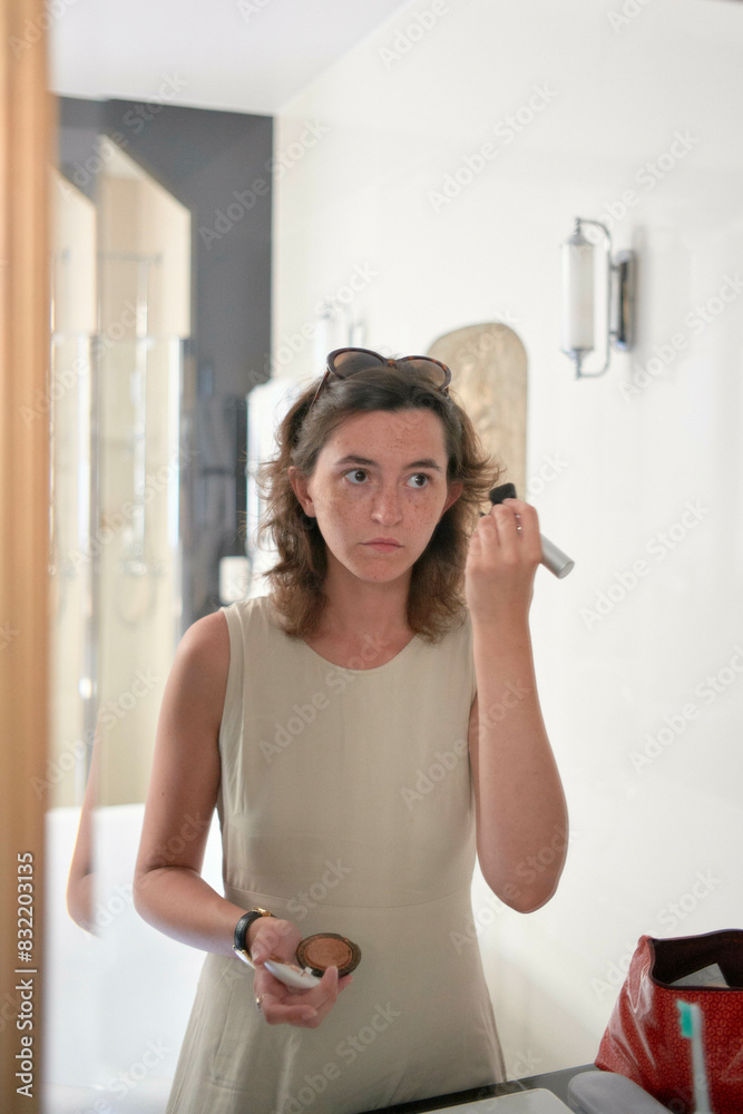 A woman is standing in front of a mirror holding a perfume bottle, preparing to spray it. She appears focused, with a neutral expression. A woman is standing in front of a mirror holding a perfume bottle, preparing to spray it. She appears focused, with a neutral expression.