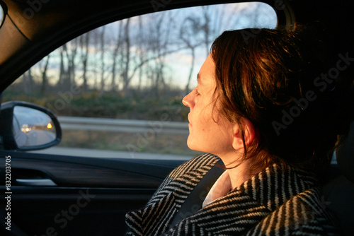 A woman is sitting inside a car looking out through the passenger window during a sunset, with gentle sunlight illuminating half of her face.