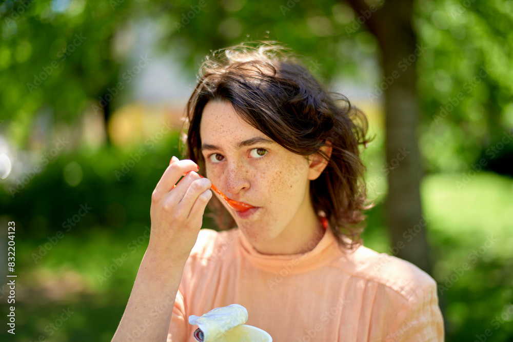 A person with freckles, wearing a peach-colored top, is eating a slice of orange, with a blurred green background. A person with freckles, wearing a peach-colored top, is eating a slice of orange, with a blurred green background.