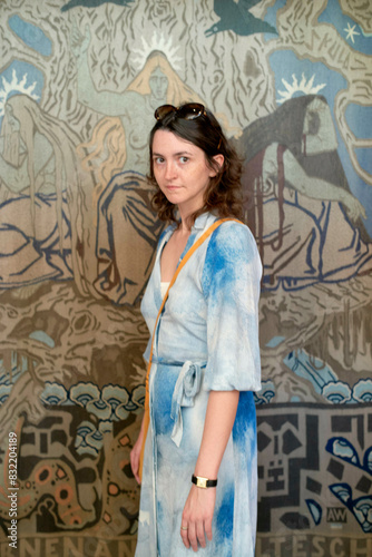 A woman stands in front of a blue-patterned wall tapestry, wearing a blue dress that blends with the background, accessorized with a shoulder bag and sunglasses. photo