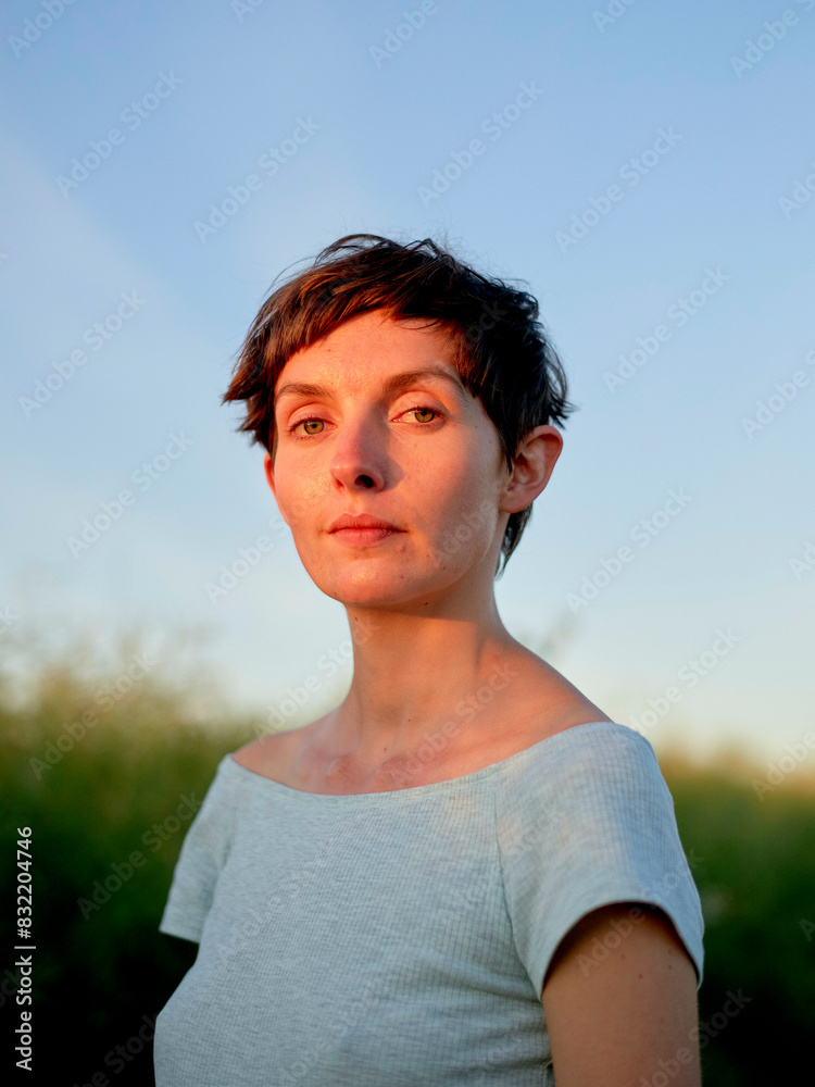 A woman with short hair stands outdoors during golden hour, her gaze directed slightly off-camera. A woman with short hair stands outdoors during golden hour, her gaze directed slightly off-camera.