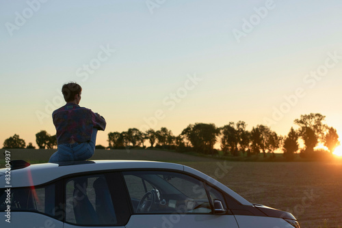 A young woman sits on top of a car, gazing at a sunset in a countryside landscape. photo