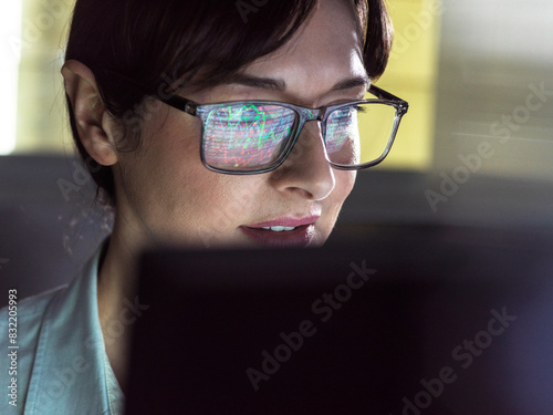 A focused individual wearing glasses with computer code reflection in the lenses is intently looking at a computer screen. photo