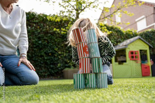 A child plays with a stack of large numbered blocks on a grass lawn, with a partially visible adult sitting nearby and a colorful playhouse in the background.