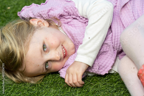 A young child with blonde hair lies on the grass, resting her head on her arm and smiling at the camera.