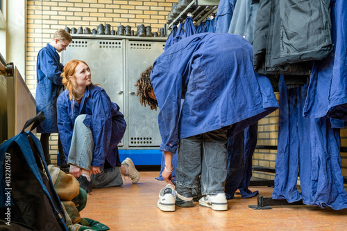Workers in protective overalls are in a locker room, with one squatting and another standing amidst lockers and hanging work garments.