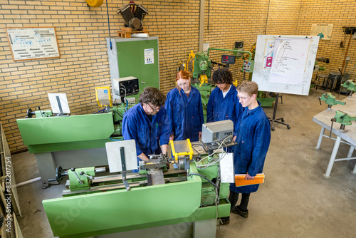 Four individuals in blue work attire are operating green industrial machinery in a workshop setting, collaborating on a technical task. photo