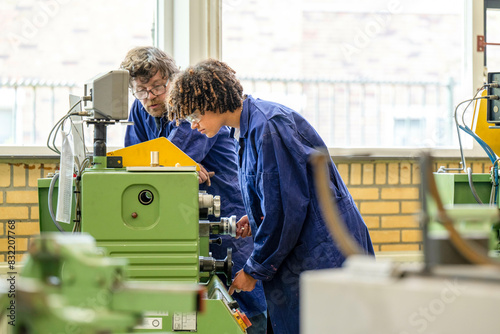 Two individuals are working attentively on a green industrial machine in a workshop setting, both wearing blue work overalls. photo