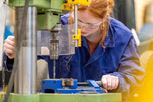 A focused individual in safety glasses and a blue work coat operates a green vertical drilling machine in an industrial setting. photo