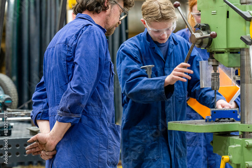 Two individuals wearing blue coveralls are working on a piece of machinery in a workshop environment, focusing intently on the task at hand. photo