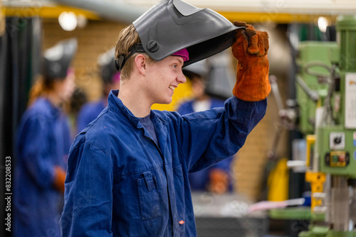A smiling young man wearing a blue work uniform is lifting his welding mask with a gloved hand in an industrial setting. photo