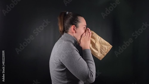 Portrait of a young woman breathing into a paper bag to calm herself during a panic attack on a black background. The struggle with anxiety and the methods used to find relief in moments of distress photo