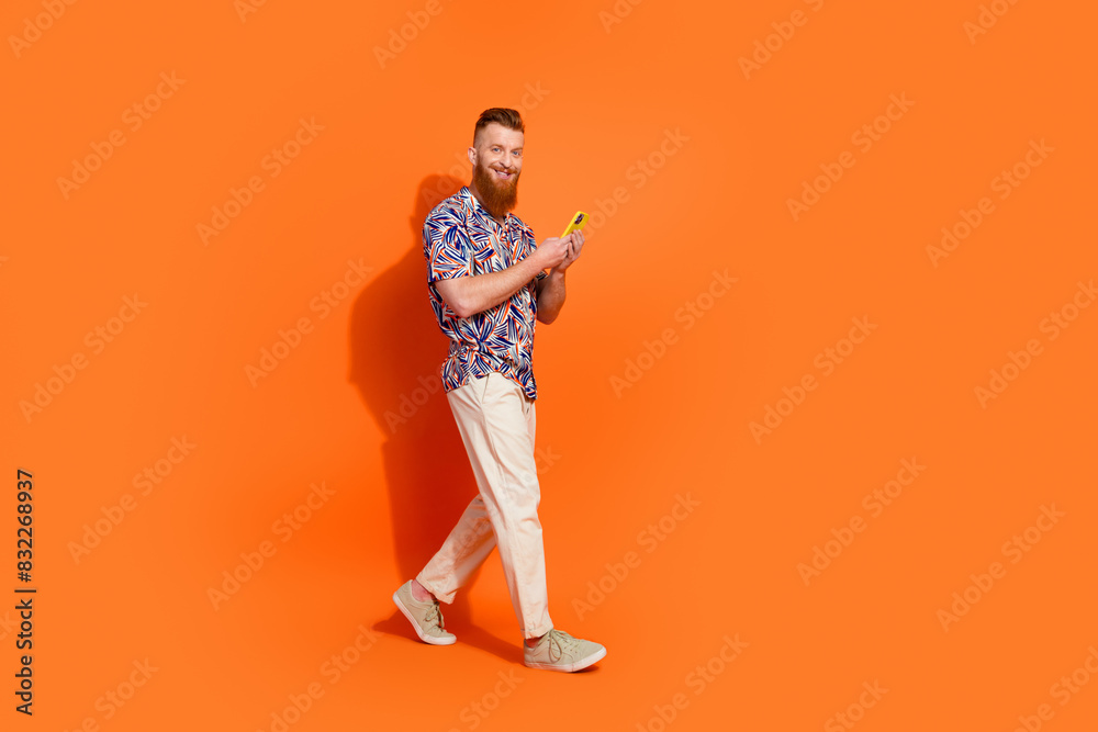 Full length photo of cute pretty guy dressed print shirt typing gadget walking empty space isolated orange color background Full length photo of cute pretty guy dressed print shirt typing gadget walking empty space isolated orange color background