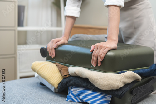 Full suitcase concept, hand of asian young woman, girl packing, preparing luggage case, luggage of clothes, female unable to close bag while sitting on sofa at home, travel trip or journey voyage. © KMPZZZ