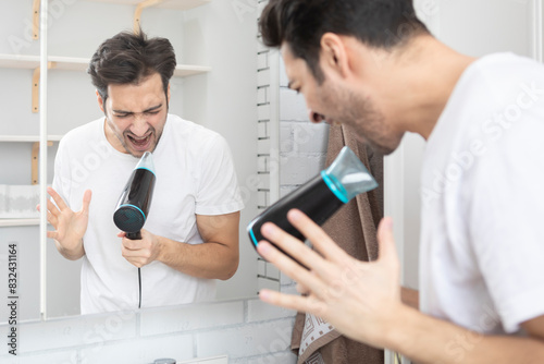 Young handsome man standing in front of the mirror in the bathroom and holding hair dryer and singing a song. Morning routine and making hair style concept