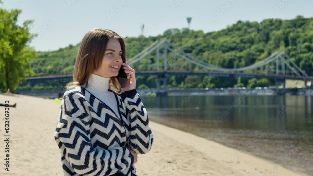 Online business in a free minute while walking. A young woman in a striped jacket talks on the phone on the bank of a city river. Online business in a free minute while walking. A young woman in a striped jacket talks on the phone on the bank of a city river.