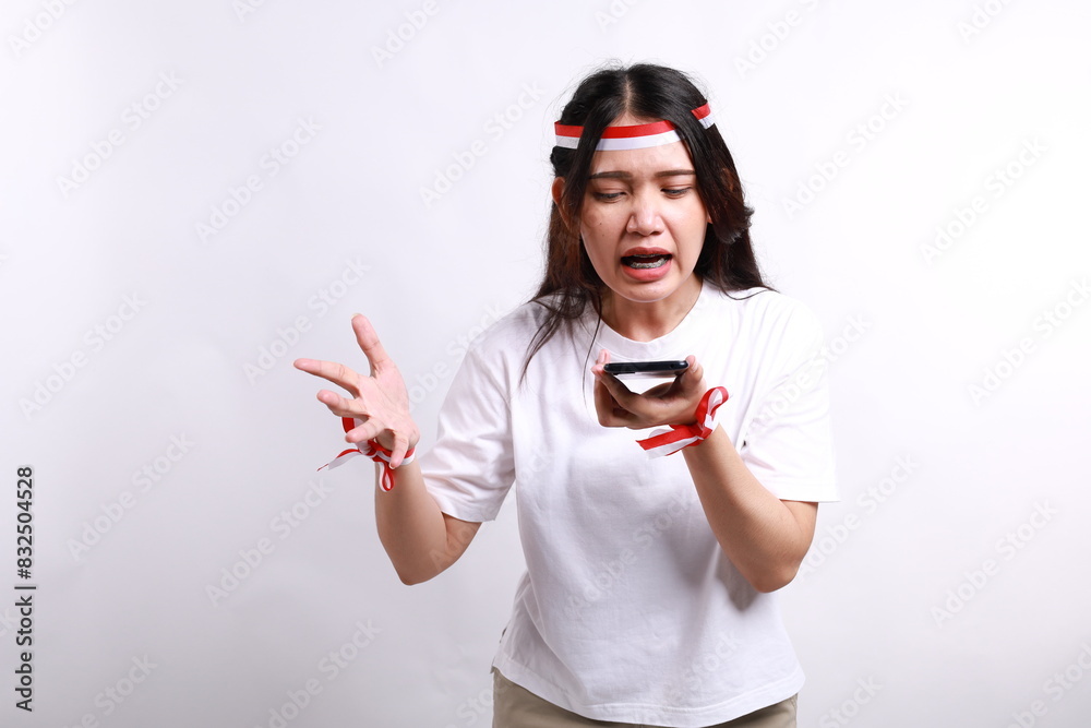 Angry young Asian woman with red white flag ribbon shouting loudly at mobile phone isolated over white background. Celebrate Indonesian independence day on 17 August Angry young Asian woman with red white flag ribbon shouting loudly at mobile phone isolated over white background. Celebrate Indonesian independence day on 17 August