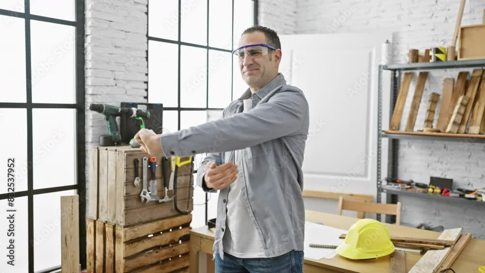 A handsome hispanic man stretching in a modern carpentry workshop with power tools and safety equipment around.