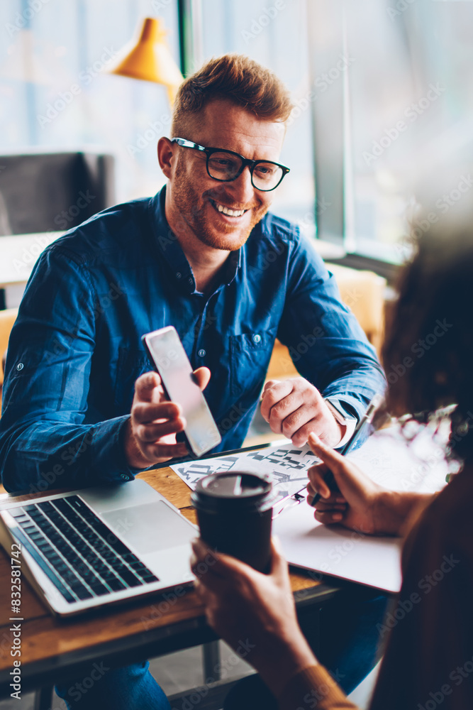 Cheerful student in red hair showing funny notification on smartphone to friend during break.Positive architect with eyewear laughing holding telephone in hand sitting at desktop with laptop Cheerful student in red hair showing funny notification on smartphone to friend during break.Positive architect with eyewear laughing holding telephone in hand sitting at desktop with laptop